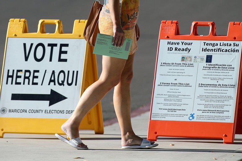 An Arizona voter carries her ballot to a polling place, walking by a "VOTE HERE" sign and a sign that tells voters to "Have Your ID Ready."